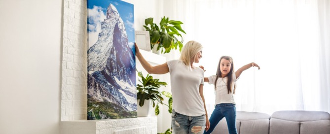 Woman and daughter hanging painting in brightly-lit room.