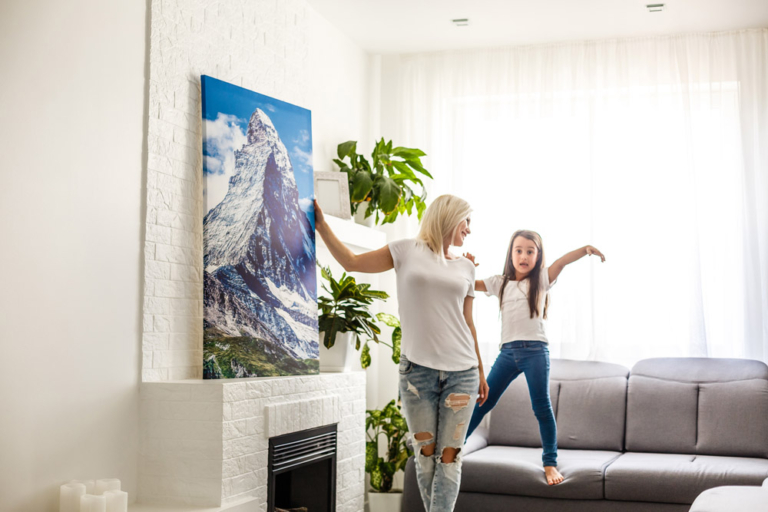 Woman and daughter hanging painting in brightly-lit room.